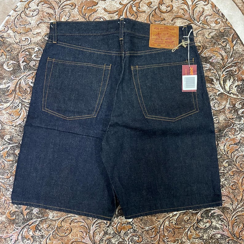 May include: Dark wash denim shorts with classic five-pocket styling. Features include a leather patch and a hang tag. The shorts have a button closure and a zip fly. The stitching is in a contrasting gold color.