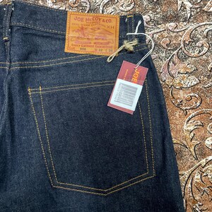 May include: Dark blue denim jeans with yellow stitching. The back pocket is visible, along with a leather patch that reads "JOE McCOY & CO." and a red tag with the brand name. The jeans are size W32 L36.