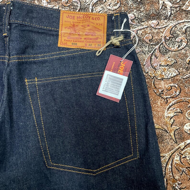 May include: Dark blue denim jeans with yellow stitching. The back pocket is visible, along with a leather patch that reads "JOE McCOY & CO." and a red tag with the brand name. The jeans are size W32 L36.