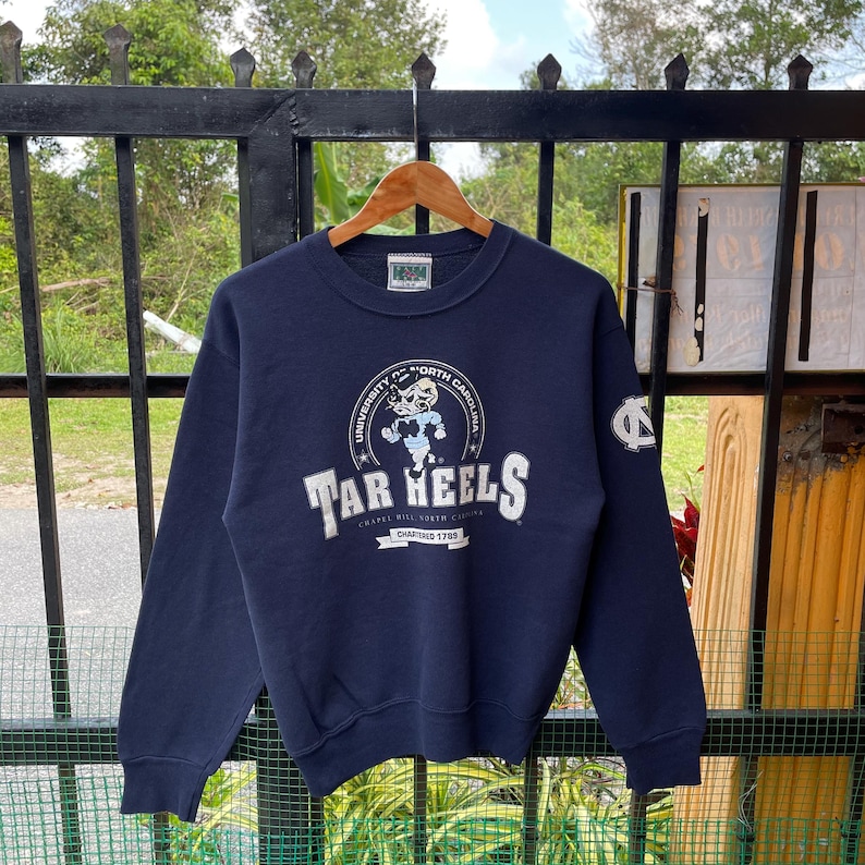 May include: Navy blue sweatshirt featuring the text "Tar Heels" in white, with a graphic of a ram and the words "University of North Carolina". The sweatshirt has a crew neck and long sleeves. The university logo is on the upper left sleeve.