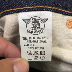 May include: Close-up of a white tag inside denim jeans. The tag features the text "The REAL McCOY'S INTERNATIONAL" and "100% COTTON" with care symbols. A copper-colored button is visible.