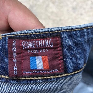 Op de afbeelding: Een close-up van een blauwe denim jeans tailleband met een rood en bordeauxrood label waarop "SOMETHING PAGEBOY" en "S/W COMPANY LIMITED" staat. Het label heeft ook een kleine Franse vlag.