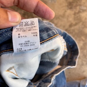 Pu&ograve; includere: Primo piano di un'etichetta di abbigliamento in denim con testo giapponese e simboli di cura. L'etichetta indica che il tessuto &egrave; 100% cotone e include le istruzioni per il lavaggio. L'etichetta riporta anche il nome del marchio e il numero di telefono.