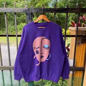 May include: A purple sweatshirt with a graphic of a stylized face. The face is primarily peach-colored with black, blue, and pink accents. The sweatshirt is hanging on a wooden hanger.