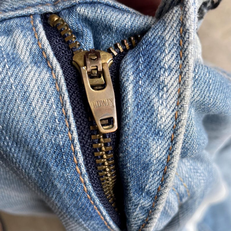 May include: Close-up of a light blue denim jeans zipper. The zipper is made of brass and features the brand name "EDWIN". The denim fabric shows signs of wear and tear, with visible stitching and a dark blue interior.