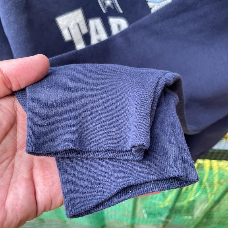 May include: Close-up of a navy blue sweatshirt sleeve. The sleeve cuff is folded over, revealing the textured fabric. White text is visible on the upper portion of the sweatshirt. The sweatshirt appears to be made of a soft, comfortable material.