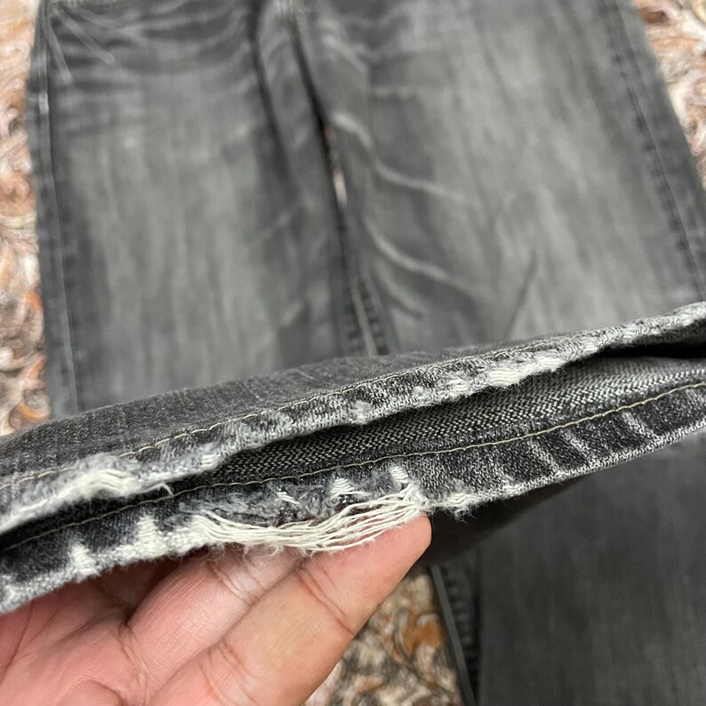 May include: Close-up of a pair of faded black denim jeans. The hem is frayed, revealing the white threads. The jeans appear worn and have a vintage aesthetic.