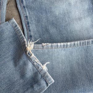 May include: Close-up of light blue denim jeans. The hem of the jeans is frayed, with visible threads and a distressed appearance. The fabric shows a classic denim texture.