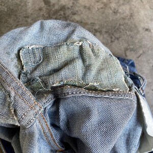 May include: Close-up of the inside waistband of blue denim jeans. The denim is faded and distressed, with visible stitching and frayed edges. The interior lining is a lighter shade of blue.