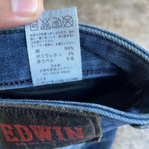 Op de afbeelding: Close-up van een blauwe denim jeans met een leren patch en een label met Japanse tekst. Het label bevat wasvoorschriften en de materiaalsamenstelling: 96% katoen, 4% polyurethaan en een leren label.
