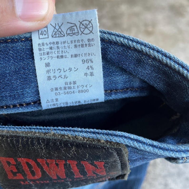 Op de afbeelding: Close-up van een blauwe denim jeans met een leren patch en een label met Japanse tekst. Het label bevat wasvoorschriften en de materiaalsamenstelling: 96% katoen, 4% polyurethaan en een leren label.