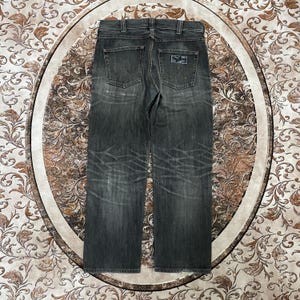 May include: Dark gray denim jeans with a classic five-pocket design. The jeans feature the Wrangler logo on the back pockets and a faded, worn appearance. The jeans have a straight leg cut.