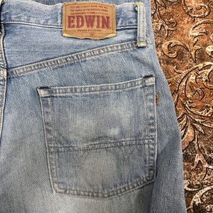 May include: Close-up of light blue denim jeans with a back pocket. A brown leather patch with the brand name "EDWIN" and text is visible. The jeans have a faded appearance.