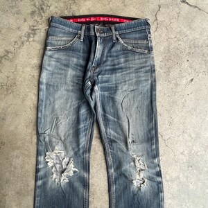 K&ouml;nnte beinhalten: Verblasste blaue Jeans mit Distressed-Details, einschlie&szlig;lich Rissen an den Knien. Die Jeans haben ein klassisches F&uuml;nf-Taschen-Design und ein rotes Bundetikett. Der Denim hat einen abgenutzten, Vintage-Look.