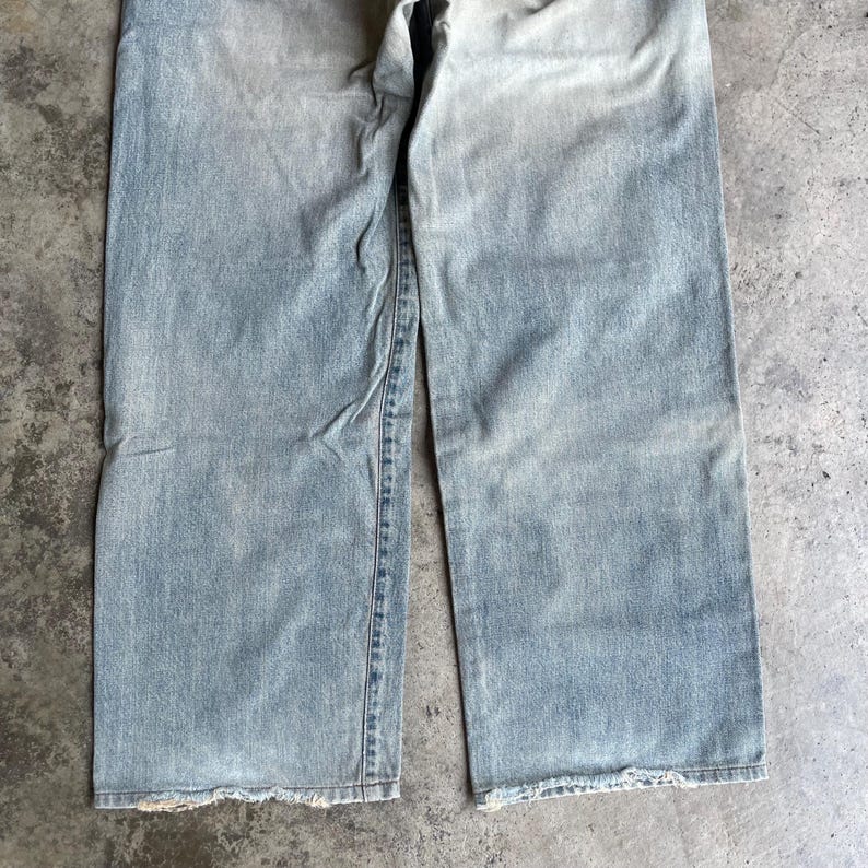 May include: Light blue denim jeans with a faded wash and a relaxed fit. The jeans have a straight leg and a frayed hem. The fabric appears worn and soft, suggesting a vintage or well-loved style. The image is a close-up of the lower portion of the pants.