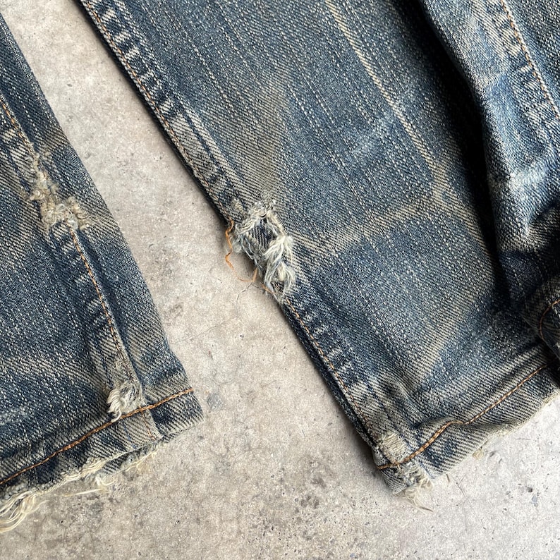 May include: Close-up of distressed blue denim jeans. The jeans show signs of wear and tear, with visible fraying and holes near the hem. The denim has a faded, vintage look, with lighter patches and subtle color variations.