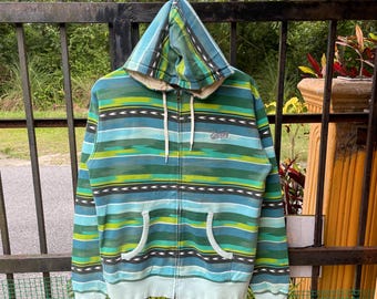 Può includere: Una felpa con cappuccio con zip a righe blu, verdi e gialle. La felpa con cappuccio ha un cappuccio con coulisse, due tasche anteriori e un piccolo logo sul petto. L'orlo e i polsini sono di un azzurro chiaro.