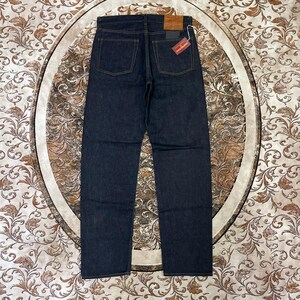 May include: Dark wash denim jeans with a straight leg cut. The jeans have classic five-pocket styling, with a brown leather patch and a red and white tag on the back. The stitching is a contrasting gold color.