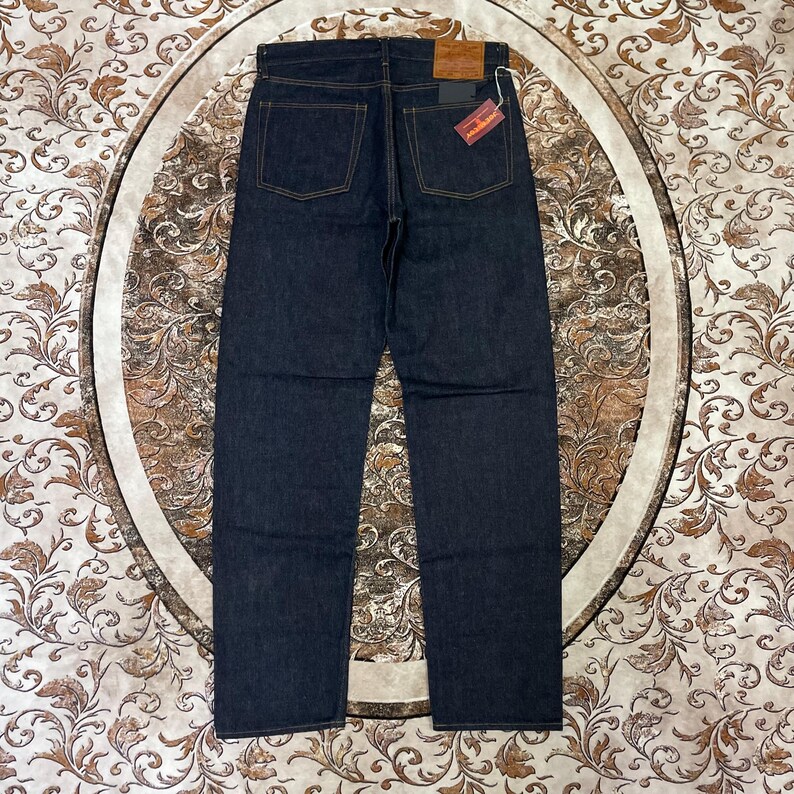 May include: Dark wash denim jeans with a straight leg cut. The jeans have classic five-pocket styling, with a brown leather patch and a red and white tag on the back. The stitching is a contrasting gold color.