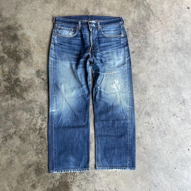May include: Faded blue denim jeans with a classic five-pocket design. The jeans show signs of wear and tear, with distressed areas on the front. The jeans have a straight leg cut and a button and zip closure.