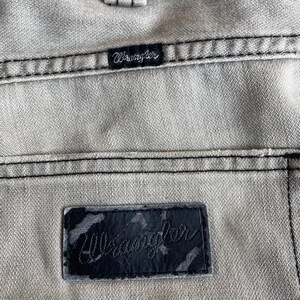 K&ouml;nnte beinhalten: Nahaufnahme von hellgrauem Denim-Stoff mit schwarzen N&auml;hten. Das Bild zeigt zwei Wrangler-Markenetiketten, eines klein und eines gr&ouml;&szlig;er, beide mit dem Markennamen in Schreibschrift.