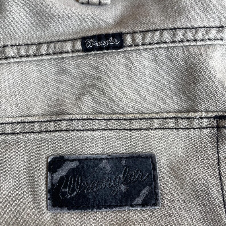 K&ouml;nnte beinhalten: Nahaufnahme von hellgrauem Denim-Stoff mit schwarzen N&auml;hten. Das Bild zeigt zwei Wrangler-Markenetiketten, eines klein und eines gr&ouml;&szlig;er, beide mit dem Markennamen in Schreibschrift.