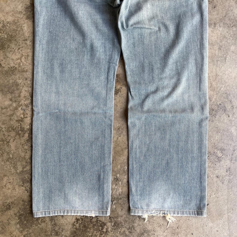 May include: Pale blue denim jeans with a faded appearance. These jeans feature a classic five-pocket design and a straight leg cut. The hem is distressed with frayed edges, giving a vintage aesthetic.