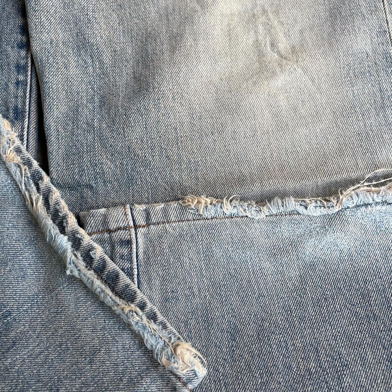 May include: Close-up of light blue denim jeans with distressed detailing. The image highlights the frayed edges and seams of the fabric, showcasing the texture and wear of the garment. The denim has a faded appearance.