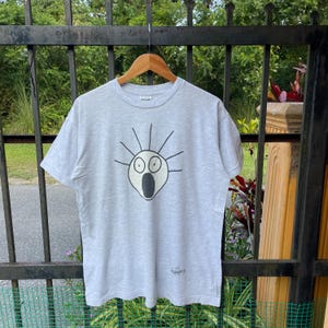 May include: A pale grey t-shirt with a cartoon face design in black and white. The design features large eyes and an open mouth. The t-shirt is hanging on a wooden hanger against a metal fence.