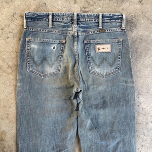 May include: Faded blue denim jeans with the Wrangler logo visible on the back pockets. The jeans show signs of wear and tear, with a distressed look. The back pockets have the classic "W" stitching.