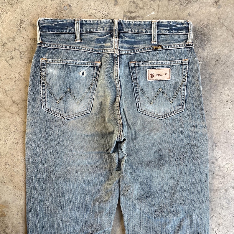 May include: Faded blue denim jeans with the Wrangler logo visible on the back pockets. The jeans show signs of wear and tear, with a distressed look. The back pockets have the classic "W" stitching.