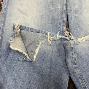 May include: Close-up of light blue denim jeans with distressed details. The jeans feature a large, ripped section on the knee, with frayed edges and a visible inner layer of fabric. The denim has a faded appearance.