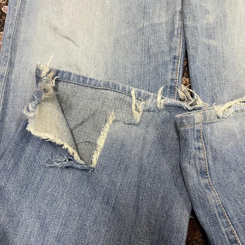 May include: Close-up of light blue denim jeans with distressed details. The jeans feature a large, ripped section on the knee, with frayed edges and a visible inner layer of fabric. The denim has a faded appearance.