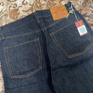 May include: Dark blue denim jeans with yellow stitching. The back pockets are square. A brown leather tag reads "JOE McCOY & CO." and a red tag with a barcode is attached. The jeans are folded.