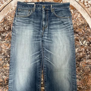 May include: A pair of blue denim jeans featuring a faded wash, a classic five-pocket design with metal rivets, a button fly closure, and visible whiskering patterns across the upper thigh area, showcasing a worn-in texture and straight leg cut.