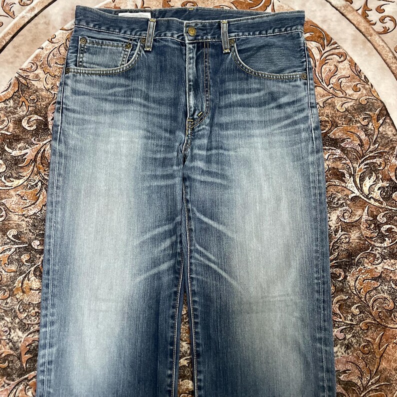 May include: A pair of blue denim jeans featuring a faded wash, a classic five-pocket design with metal rivets, a button fly closure, and visible whiskering patterns across the upper thigh area, showcasing a worn-in texture and straight leg cut.