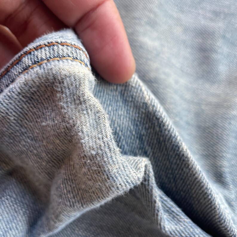 May include: Close-up of light blue denim fabric with visible texture and brown stitching along the edge. The fabric is folded, revealing the inside texture of the denim. The image highlights the details of the material.