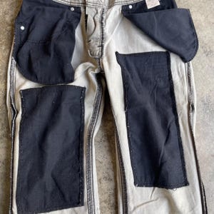 K&ouml;nnte beinhalten: Eine helle Jeanshose mit gro&szlig;en, dunklen Flicken auf der Vorder- und R&uuml;ckseite. Die Taschen sind sichtbar, und die N&auml;hte sind in einer kontrastierenden dunklen Farbe gehalten. Die Hose hat einen Knopf- und Rei&szlig;verschluss.