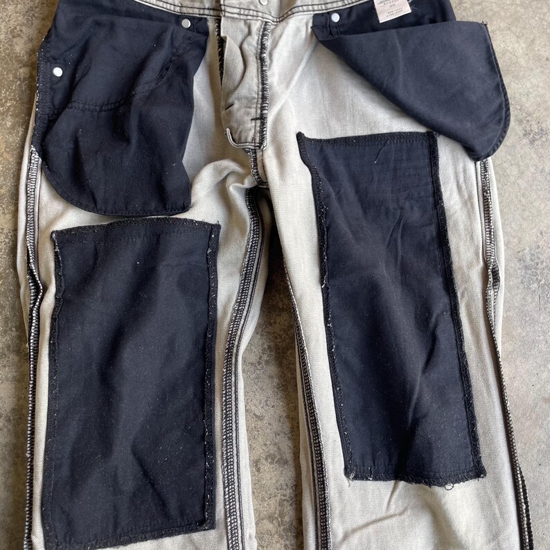 K&ouml;nnte beinhalten: Eine helle Jeanshose mit gro&szlig;en, dunklen Flicken auf der Vorder- und R&uuml;ckseite. Die Taschen sind sichtbar, und die N&auml;hte sind in einer kontrastierenden dunklen Farbe gehalten. Die Hose hat einen Knopf- und Rei&szlig;verschluss.