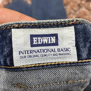Puede incluir: Primer plano de una etiqueta de vaqueros de mezclilla con el nombre de la marca "EDWIN" en un rect&aacute;ngulo azul marino. Debajo, la etiqueta dice "INTERNATIONAL BASIC OUR ORIGINAL QUALITY AND WASHING". Los vaqueros son de lavado medio.