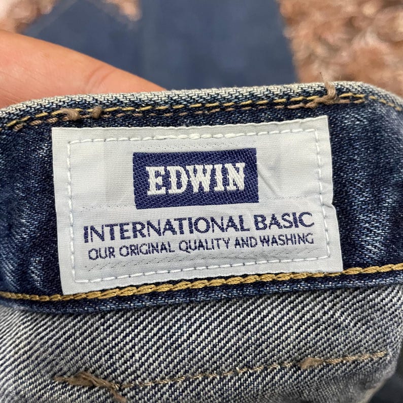 Puede incluir: Primer plano de una etiqueta de vaqueros de mezclilla con el nombre de la marca "EDWIN" en un rect&aacute;ngulo azul marino. Debajo, la etiqueta dice "INTERNATIONAL BASIC OUR ORIGINAL QUALITY AND WASHING". Los vaqueros son de lavado medio.