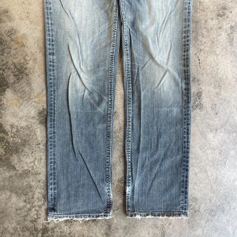 May include: Faded blue denim jeans with a straight leg cut. The jeans have a worn appearance with visible fading and distressing. The bottom hems are frayed. The jeans are made of a durable denim material.