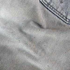May include: Close-up of light blue denim fabric with a pocket detail. The denim shows signs of wear and tear, with visible fading and discoloration. The texture of the fabric is clearly visible, highlighting the weave of the material.