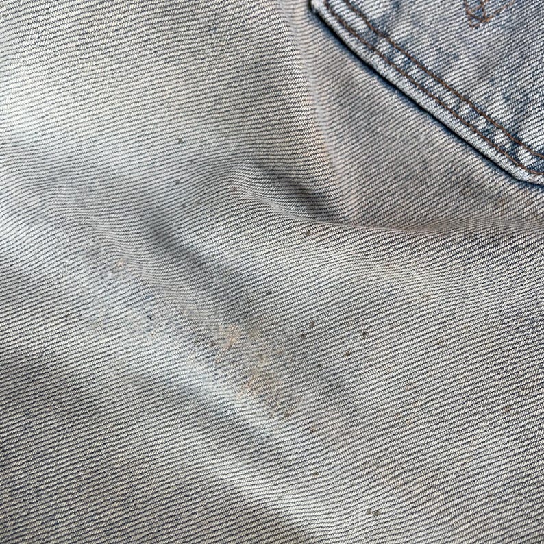 May include: Close-up of light blue denim fabric with a pocket detail. The denim shows signs of wear and tear, with visible fading and discoloration. The texture of the fabric is clearly visible, highlighting the weave of the material.