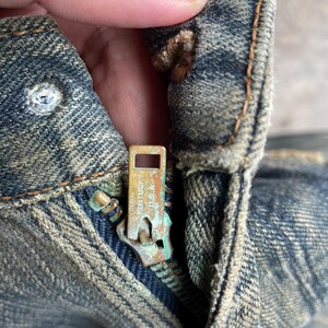 May include: Close-up of a distressed denim jeans zipper. The zipper pull is a tarnished brass color, with the text "Kentucky U.S.A." visible. The denim has a faded blue wash with visible wear and tear.