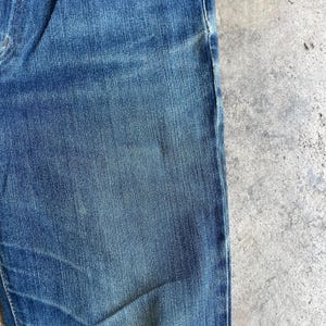 Op de afbeelding: Close-up van een blauwe spijkerbroek. De stof heeft een getextureerde weefsel met lichtere en donkerdere plekken, wat een versleten uitstraling geeft. Een deel van de broekspijp en de zoom zijn zichtbaar.