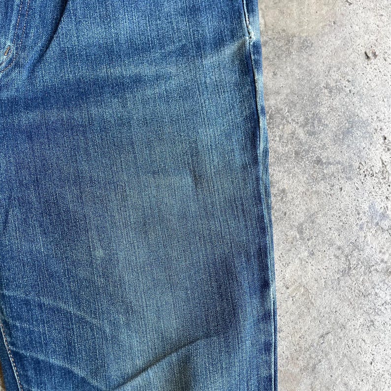 Op de afbeelding: Close-up van een blauwe spijkerbroek. De stof heeft een getextureerde weefsel met lichtere en donkerdere plekken, wat een versleten uitstraling geeft. Een deel van de broekspijp en de zoom zijn zichtbaar.