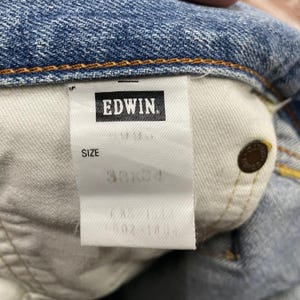 May include: Close-up of the inside label of a pair of blue denim jeans. The label reads "EDWIN" in a black rectangle. The size is listed as 30x34. A copper-colored button is visible on the jeans.