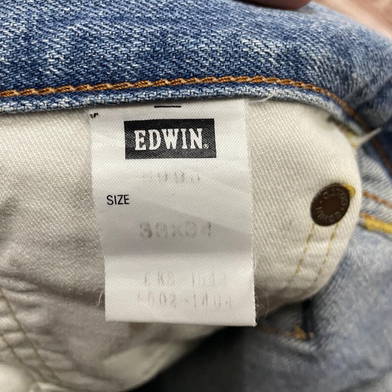 May include: Close-up of the inside label of a pair of blue denim jeans. The label reads "EDWIN" in a black rectangle. The size is listed as 30x34. A copper-colored button is visible on the jeans.