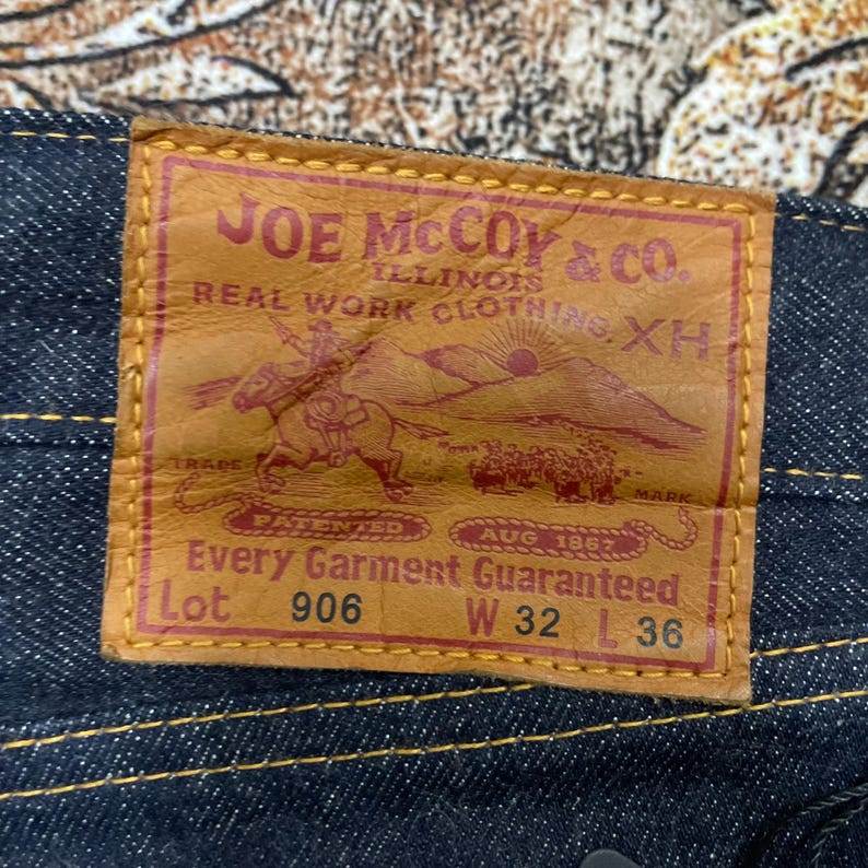 May include: Close-up of a brown leather label on dark blue denim jeans. The label reads "JOE McCOY & Co. ILLINOIS REAL WORK CLOTHING XH" with a graphic of a cowboy on a horse. The label also indicates the jeans are size W32 L36.